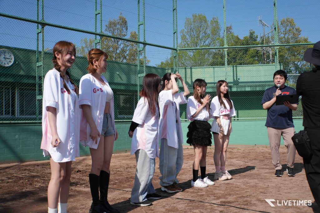Group of young women in white uniforms standing on a dirt baseball field, a man with a microphone addressing them near a green chain-link fence on a sunny day.
