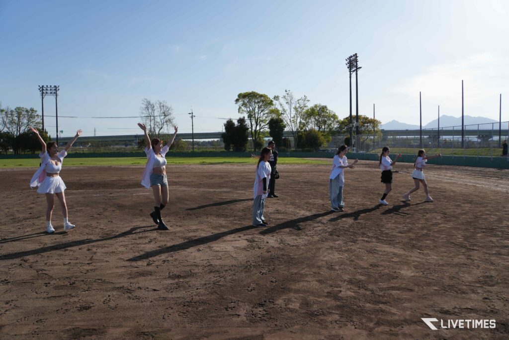 Group of young women in white outfits dancing on a dusty baseball field, with floodlights, trees, and distant mountains in the background.
