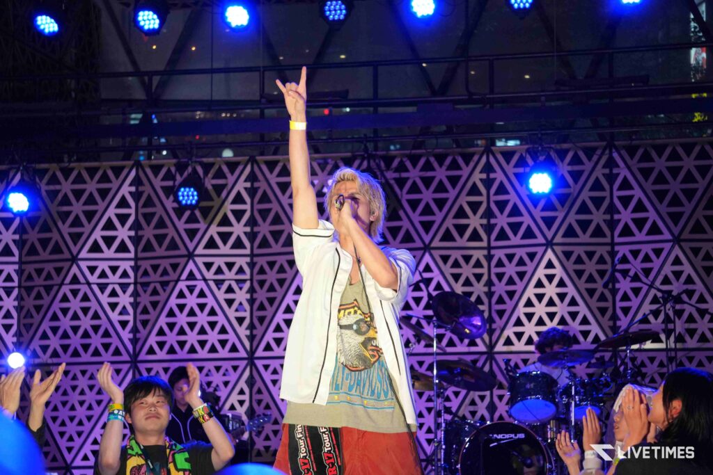 Performer on stage raising his right hand in a rock gesture, microphone in left hand, colorful stage lights and cheering audience nearby.
