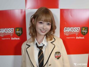 Smiling young woman in a beige blazer and striped tie posing in front of a red-and-white backdrop with logos.