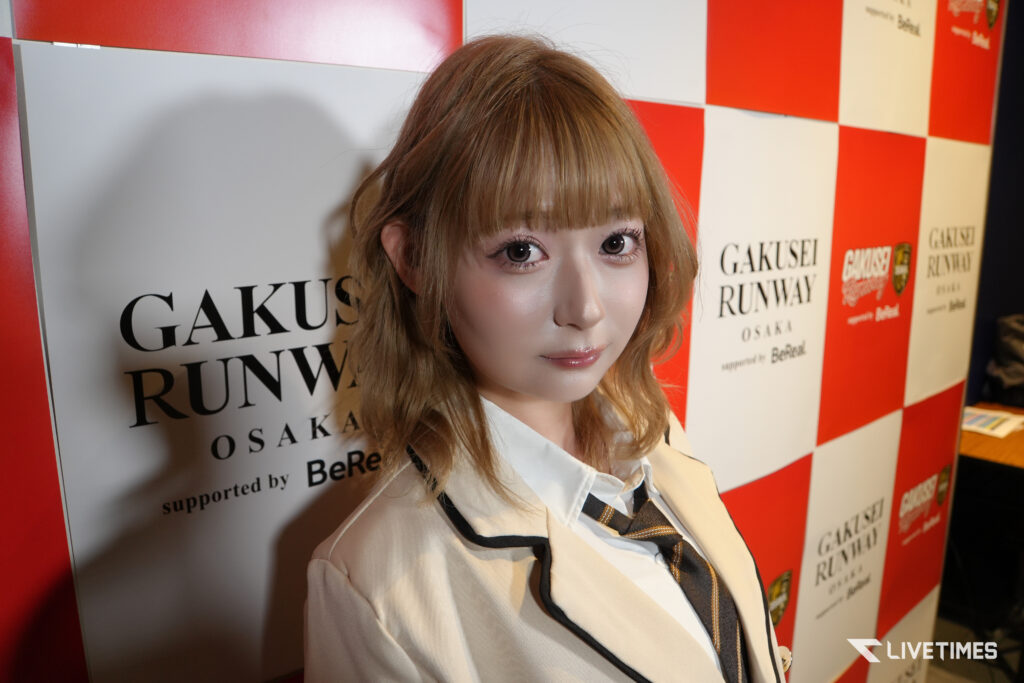 Young woman in a beige uniform with a striped tie posing in front of a red-and-white checkered backdrop that reads 'GAKUSEI RUNWAY OSAKA'.