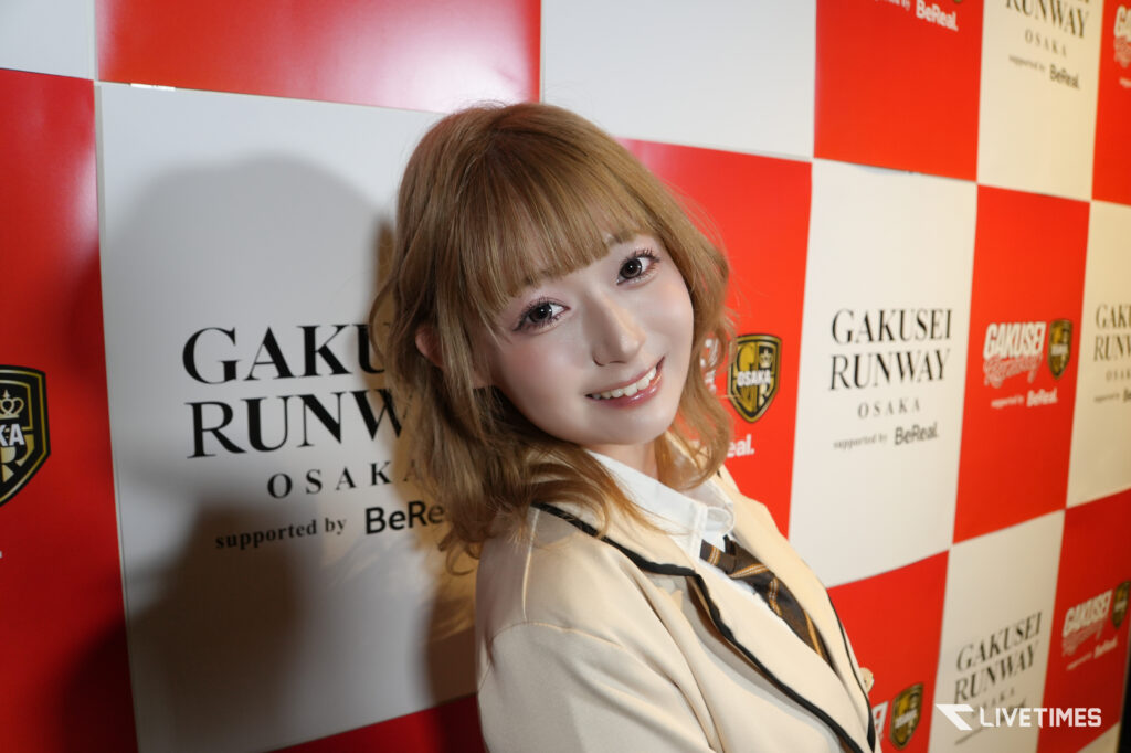 Young woman with light brown hair smiling at the camera in front of a red-and-white sponsor backdrop with logos.