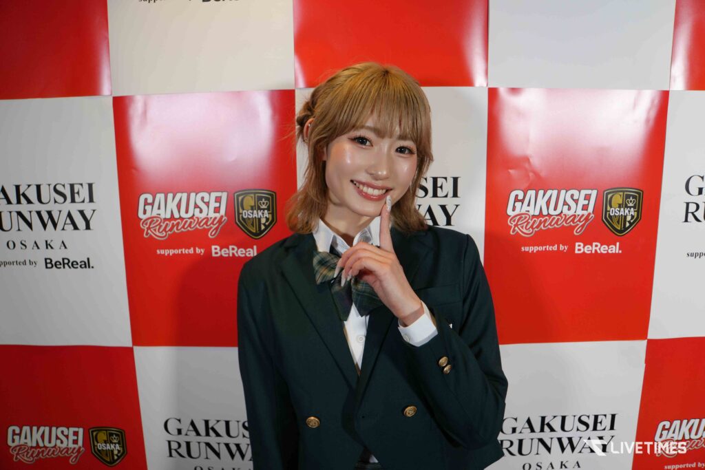Young woman in a school blazer posing with a finger to her lips against a red-and-white checkered backdrop with logos (Gakusei Runway Osaka).