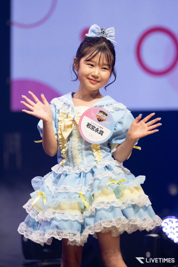 Young girl in a light blue frilly dress waves on stage, with a round contestant badge pinned to her chest and a tiara on her head.