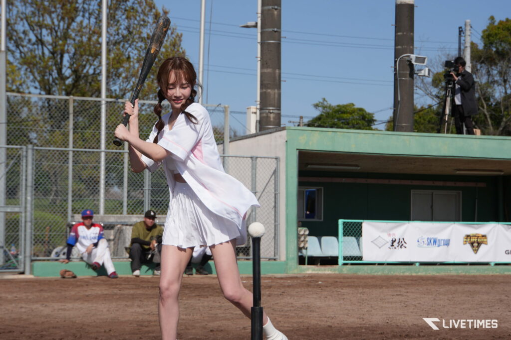Young girl in a white baseball uniform swings a bat during batting practice on a dirt field, with a photographer on a platform in the background.