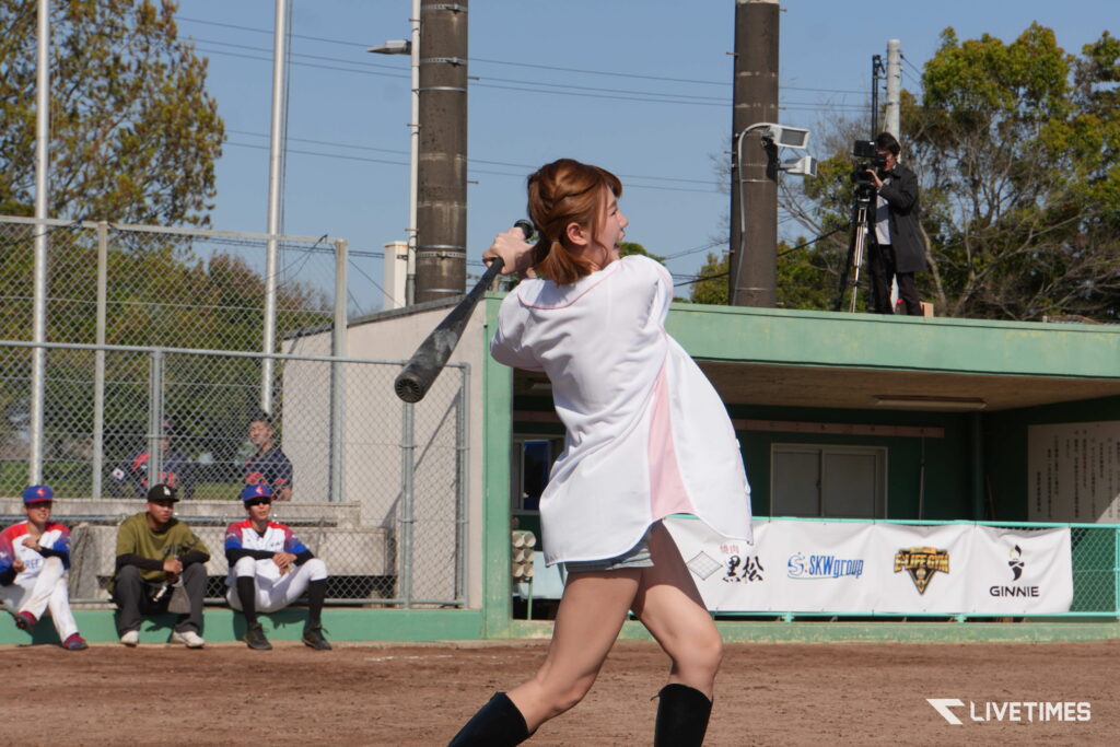 Female performer in white sings into a microphone on a baseball field with a cameraman filming from behind.