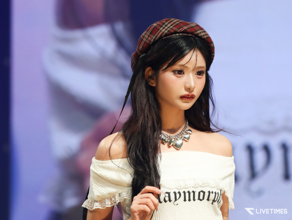 Young woman with long dark hair wearing a red plaid beret and off-the-shoulder white top on stage.
