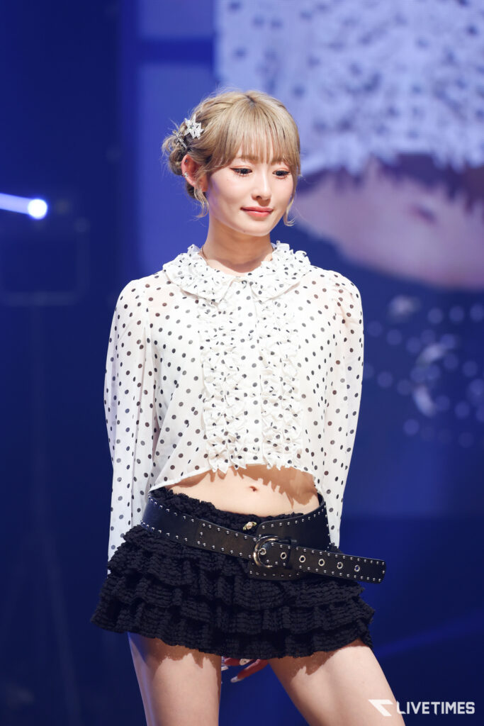 Female performer on stage wearing a white polka-dot blouse with ruffles and a black layered skirt, under blue stage lighting.