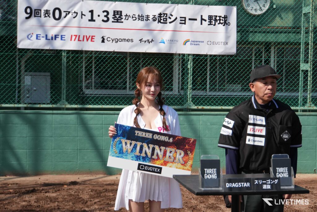 Woman in a white dress holding a large WINNER sign at a sports award ceremony, with a man in a black cap beside her and sponsor banners in the background on a green fence.