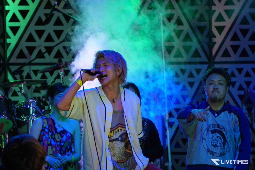Singer on stage singing into a microphone as colorful fog and geometric backdrop glow behind him