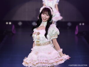 Smiling performer in a pastel pink and cream frilly dress on stage, posing for the audience.