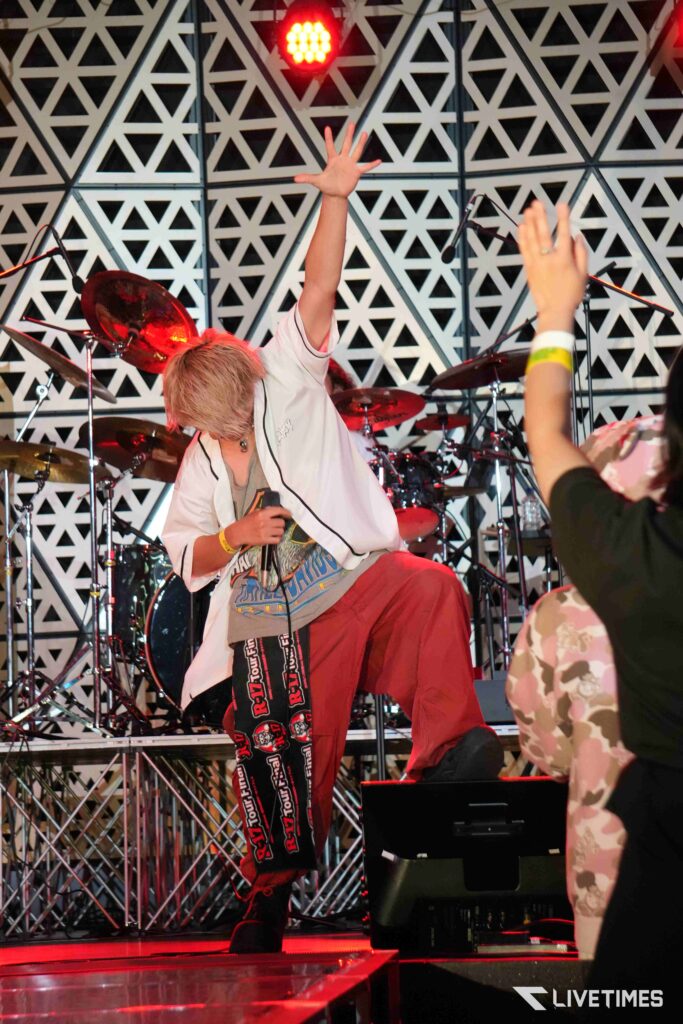Energetic musician on stage raising a hand, with drums behind and a geometric patterned wall in the background.