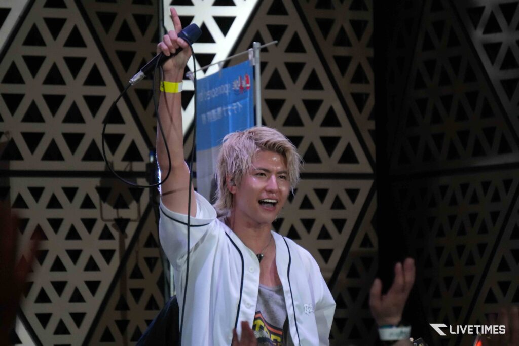 Male performer with blond hair raises a microphone on stage, wearing a white shirt and a yellow wristband.