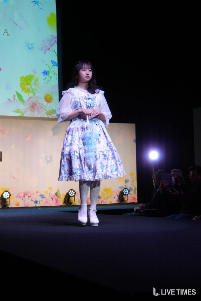 Young woman in a light blue floral dress on stage, hands clasped, with a flower-pattern backdrop and audience in the dim foreground.
