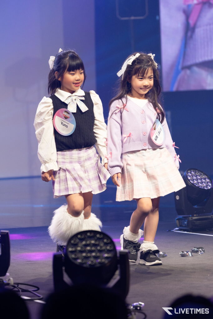Two young girls on a stage wearing pastel outfits and badges, smiling during a performance.