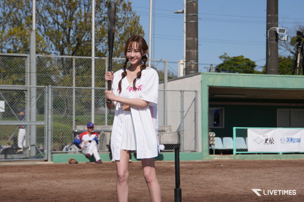 Young woman in a white baseball uniform holding a bat, standing on a dirt field with fences and dugout behind her, smiling at the camera.