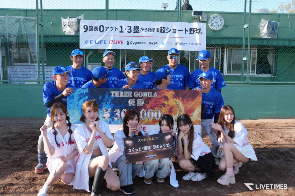 Group of baseball players in blue uniforms posing with cheerleaders and a large prize banner on a dirt field near a dugout.