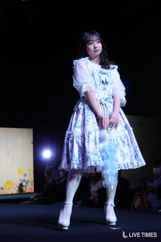 Young female performer on a dark stage wearing a light blue, floral, frilly dress with lace and bows, hands gently clasped in front during a live performance