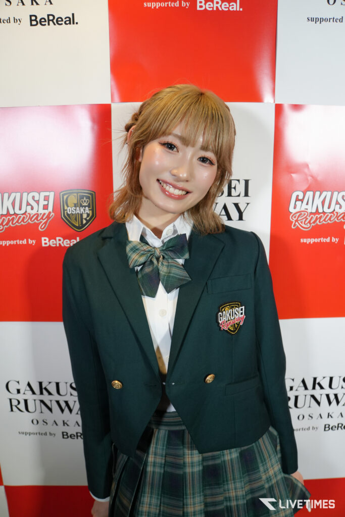 Smiling young woman in a green blazer and plaid bow, posing in front of a red-and-white sponsor backdrop (GAKUSEI RUNWAY OSAKA).