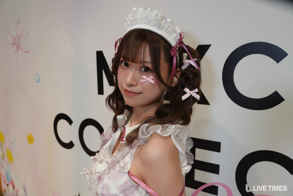 Cosplay model in a white lace maid outfit with pink ribbons, posing in front of a backdrop with large text.
