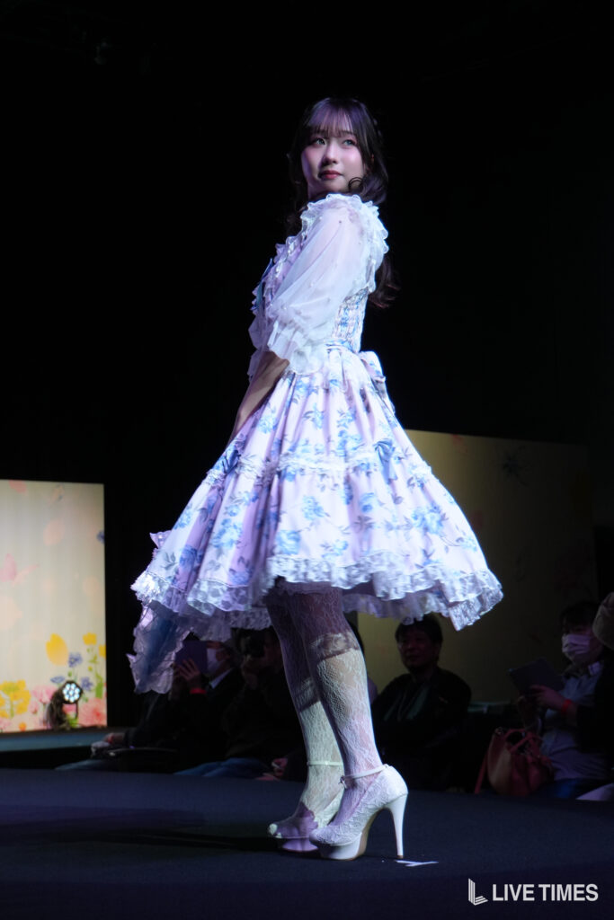 Performer on stage in a pastel blue floral dress with ruffles, white lace tights, and high heels, seen from the side.