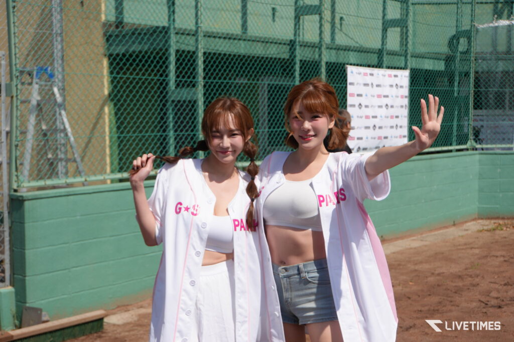 Two young women in white outfits with pink lettering pose and wave at a sunny baseball field behind a green chain-link fence.