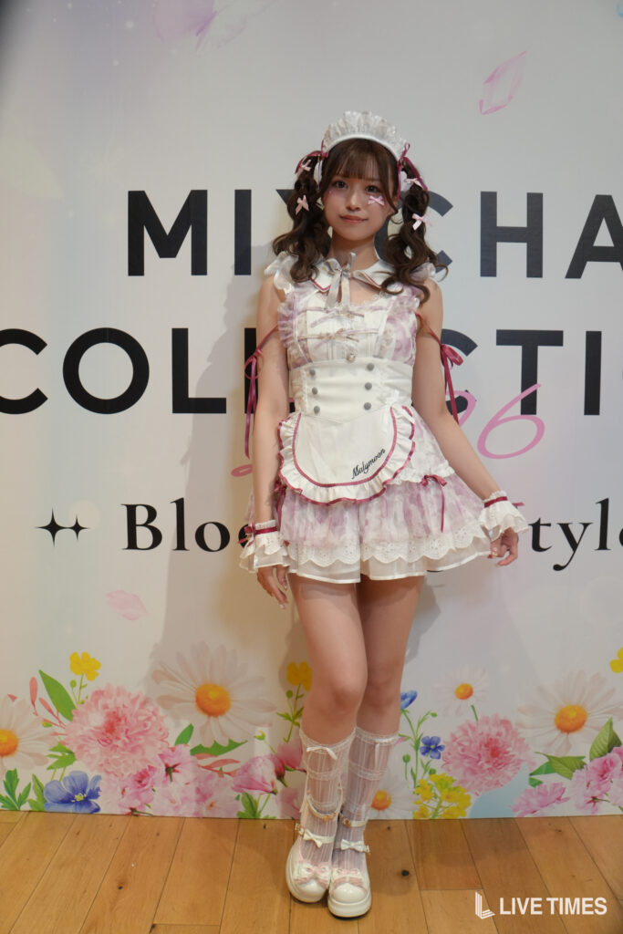 Young woman in a pink-and-white maid cosplay with a lace headpiece, posing in front of a large-letter backdrop and floral border.