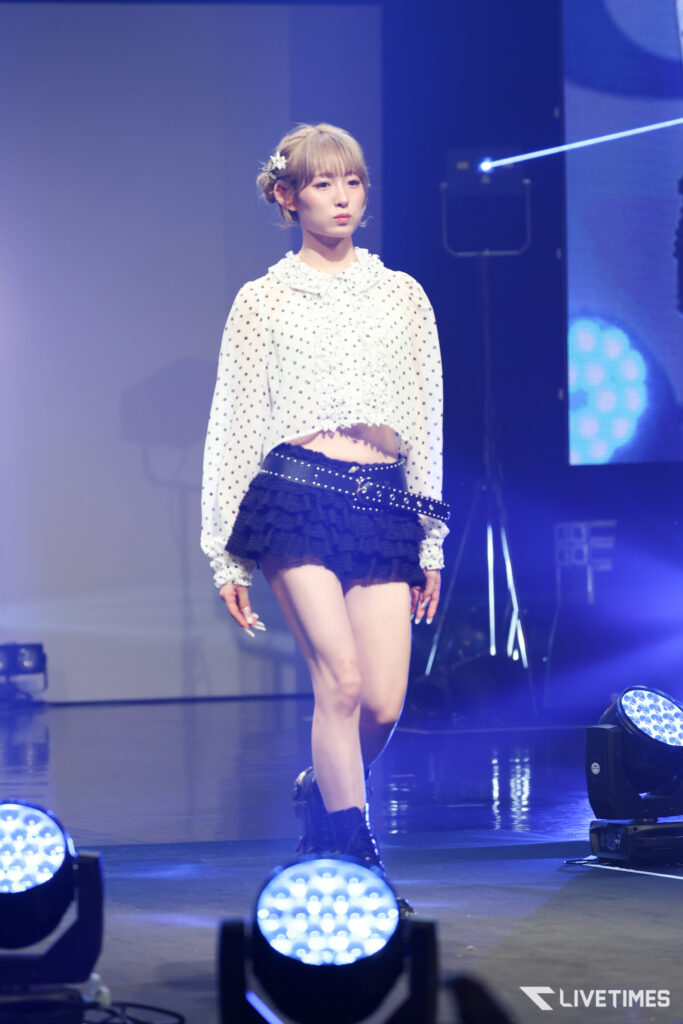 Young female performer on stage in a white polka-dot blouse and black ruffled mini skirt, with a hair accessory in place.