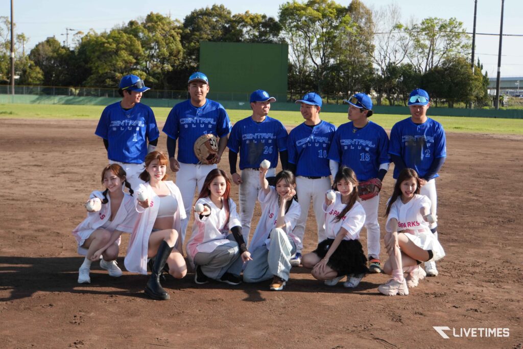 Baseball team in blue jerseys posing for a group photo on the field with smiling girls kneeling in front.