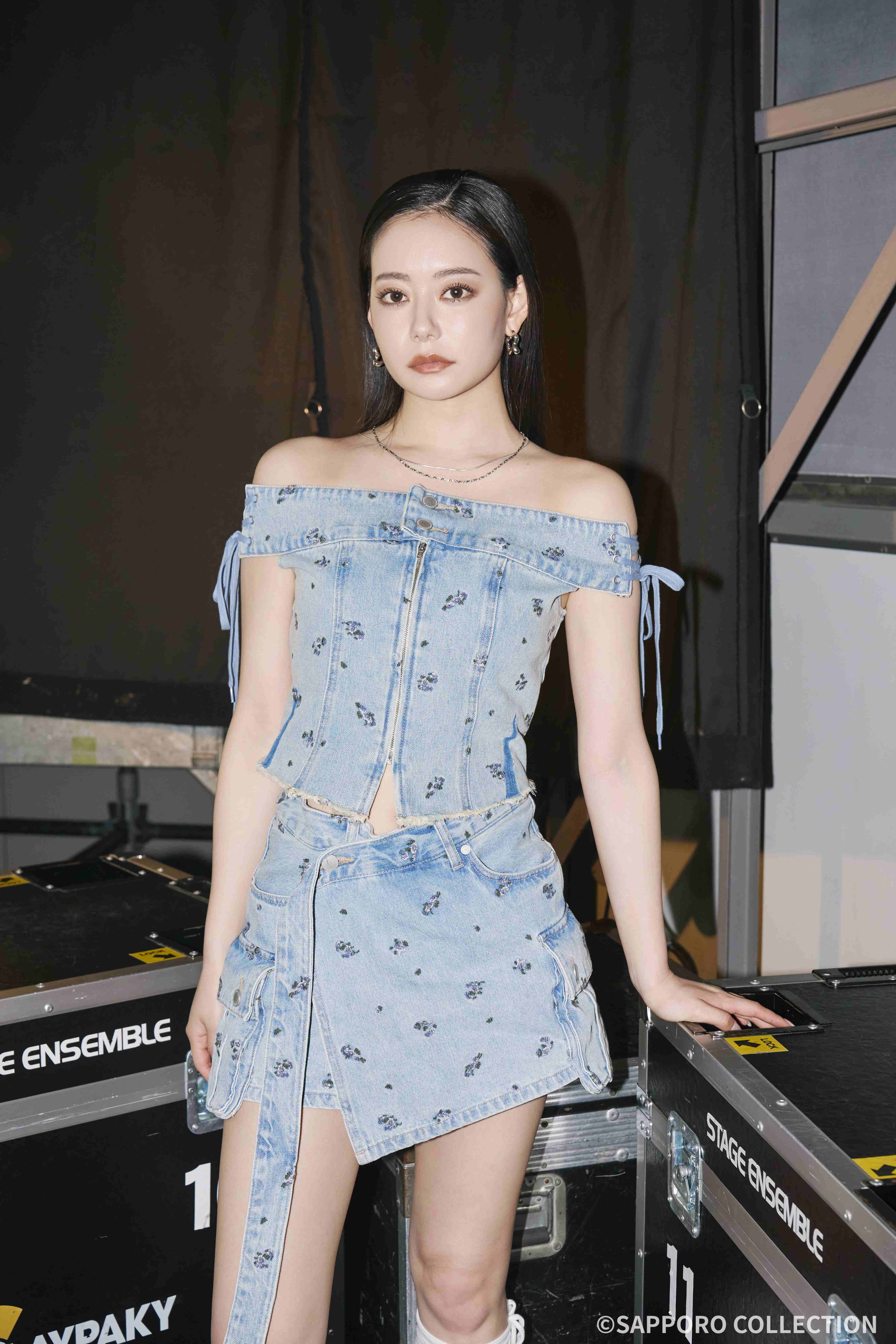 Woman in a light denim off-shoulder top and matching mini skirt with small floral print, standing beside stage equipment backstage.