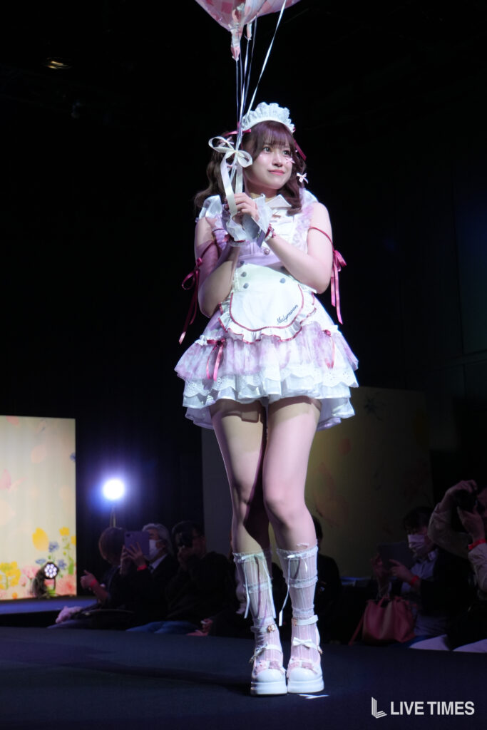 Young woman in a pink-and-white maid outfit on stage, holding pastel balloons and looking upward with a gentle smile.