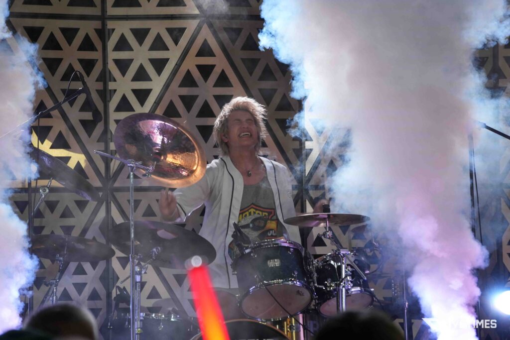 Drummer on stage amid smoke, mid-performance with sticks raised, surrounded by cymbals and a geometric triangle backdrop.