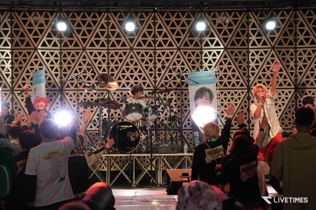 Live concert on a stage with a drummer center, a singer to the right, and an excited crowd with raised hands in front of a geometric backdrop.