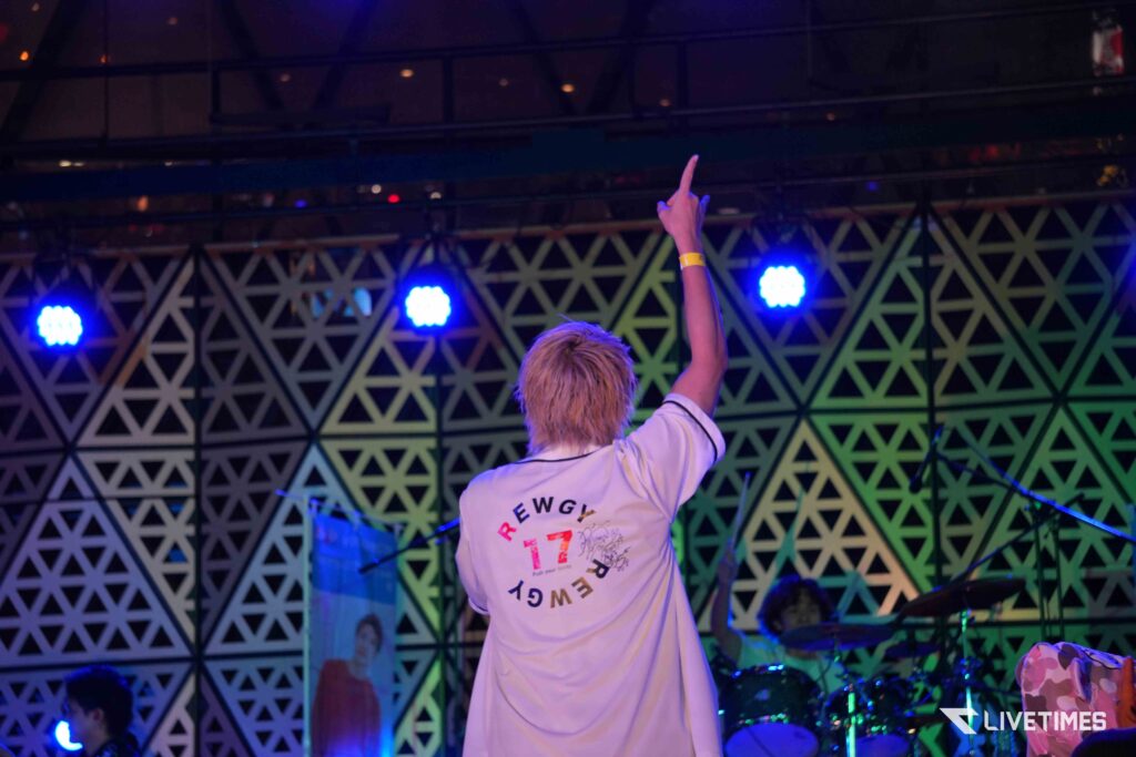 Person on stage, back to camera, raising a finger in the air while wearing a white graphic T-shirt, with a geometric backdrop and blue stage lights.