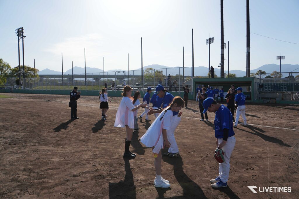 Young baseball players in blue uniforms greet each other on a dirt field, with a dugout, bleachers, and mountains in the distant background.