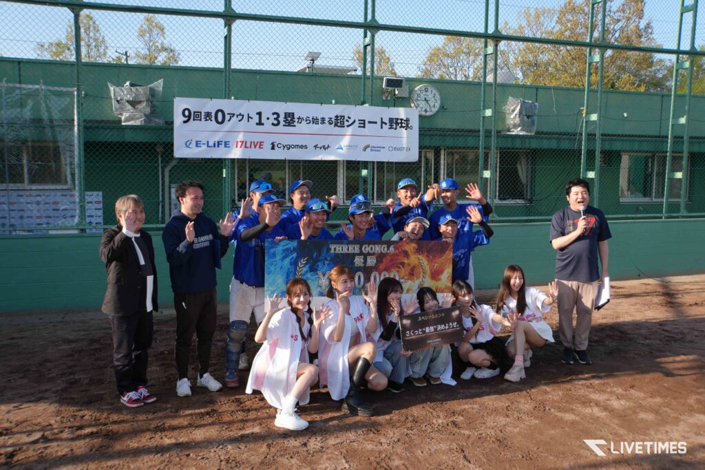 Group of players in blue uniforms and supporters posing with a banner at a baseball field, green fence in background and a clock visible above.