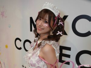Young woman in a white lace maid costume with pink ribbons posing and smiling at a backdrop with large letters.