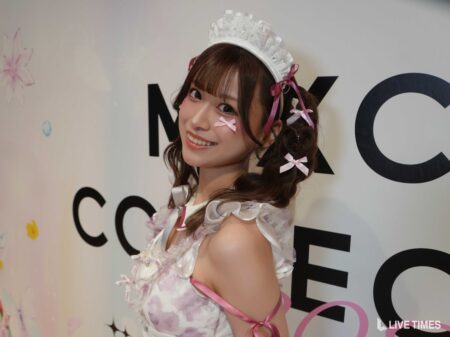 Young woman in a white lace maid costume with pink ribbons posing and smiling at a backdrop with large letters.
