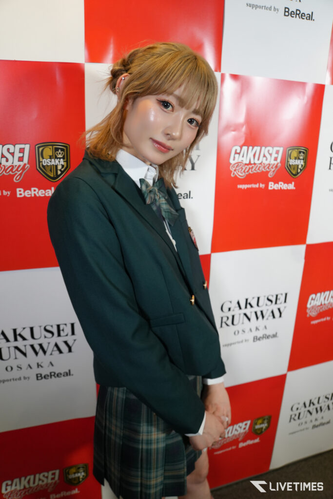 Young woman in a dark blazer and plaid skirt posing in front of a red-and-white backdrop with GAKUSEI RUNWAY Osaka branding.