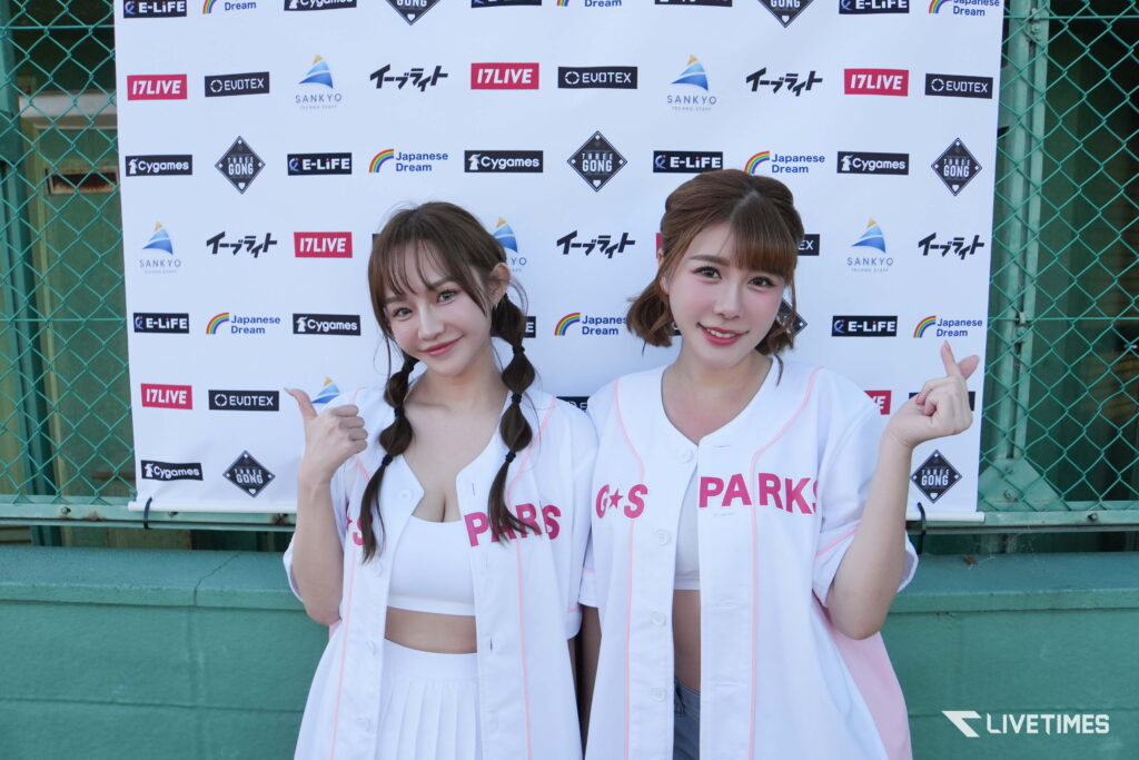 Two smiling young women in white uniforms pose in front of a sponsor banner and green chain-link fence at an outdoor event.