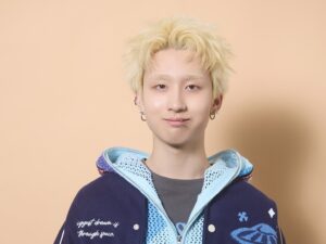 Young person with bleached blonde hair, wearing a navy varsity jacket over a light blue hoodie, facing the camera with a subtle smile and multiple ear piercings.