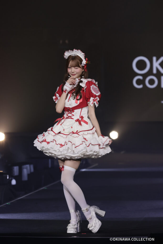 Young woman in a red and white frilly maid outfit performing on a stage, hands near her face, with stage lights in the background.