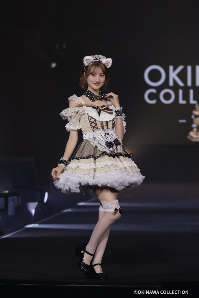 Model on a runway wearing a beige-lace lolita dress with white frills, black polka-dot accents, and cat-ear headband, smiling at the camera on stage with 'OKINAWA COLLECTION' in the background.