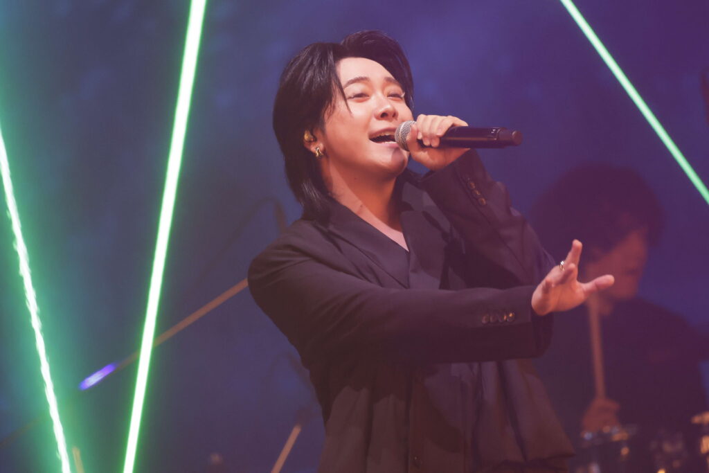 Performer singing into a handheld microphone on a dark stage with bright green laser lights in the background, wearing a black blazer.