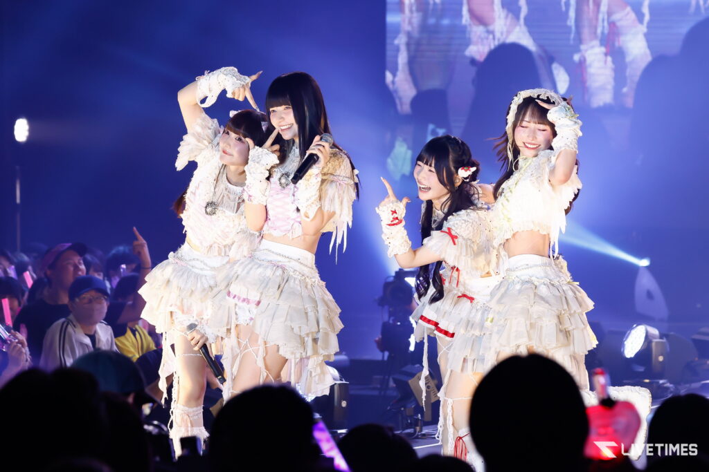 Four female performers in frilly white outfits sing and pose on a blue-lit stage, microphone in hand, engaging the cheering audience.