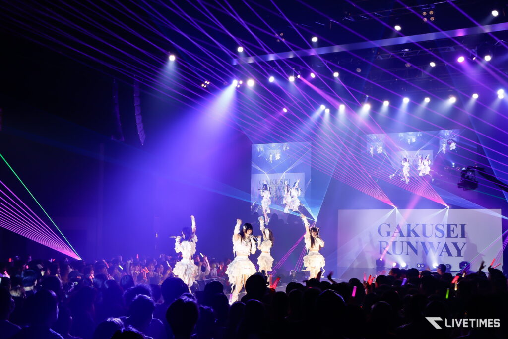 Stage concert with purple laser beams and dancers in white outfits performing on a lit, multi-screen backdrop while the cheering crowd waves glow sticks.