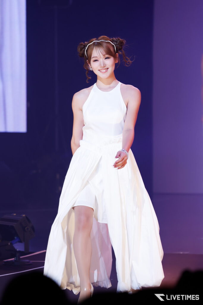 Smiling female performer in a white halter dress on stage, with a sparkly headband and two buns in her hair, under purple stage lights.