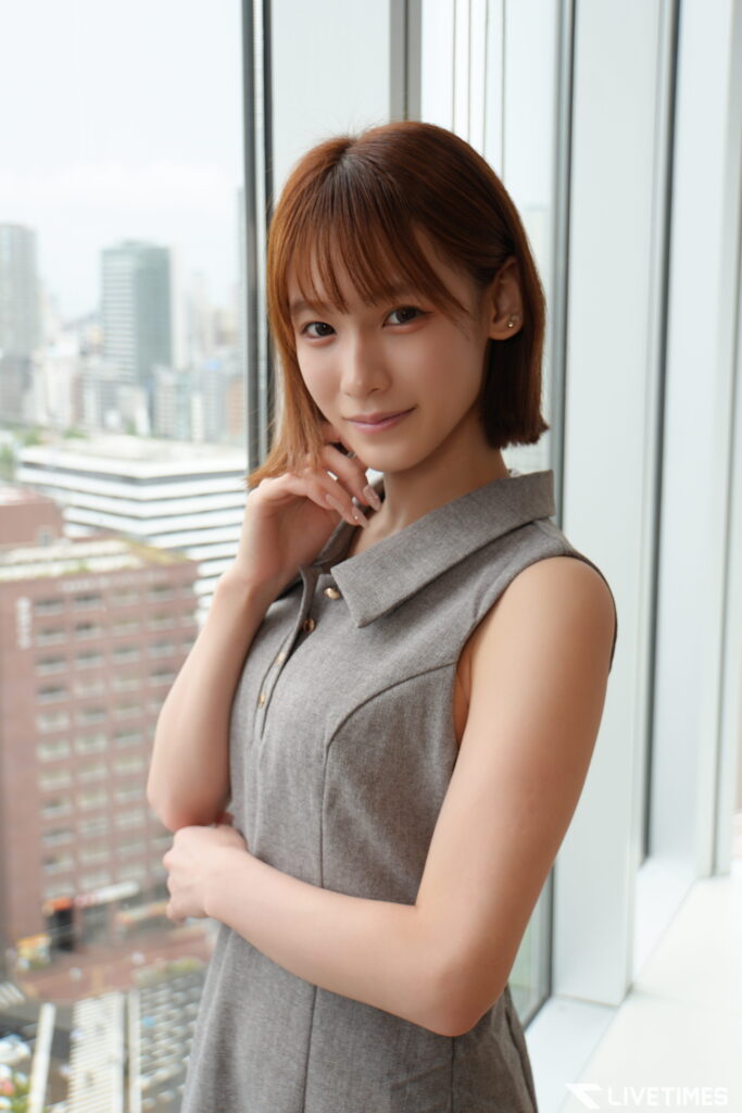 Portrait of a young woman standing by a large window with a city skyline in the background, hand resting near her chin, wearing a sleeveless gray dress