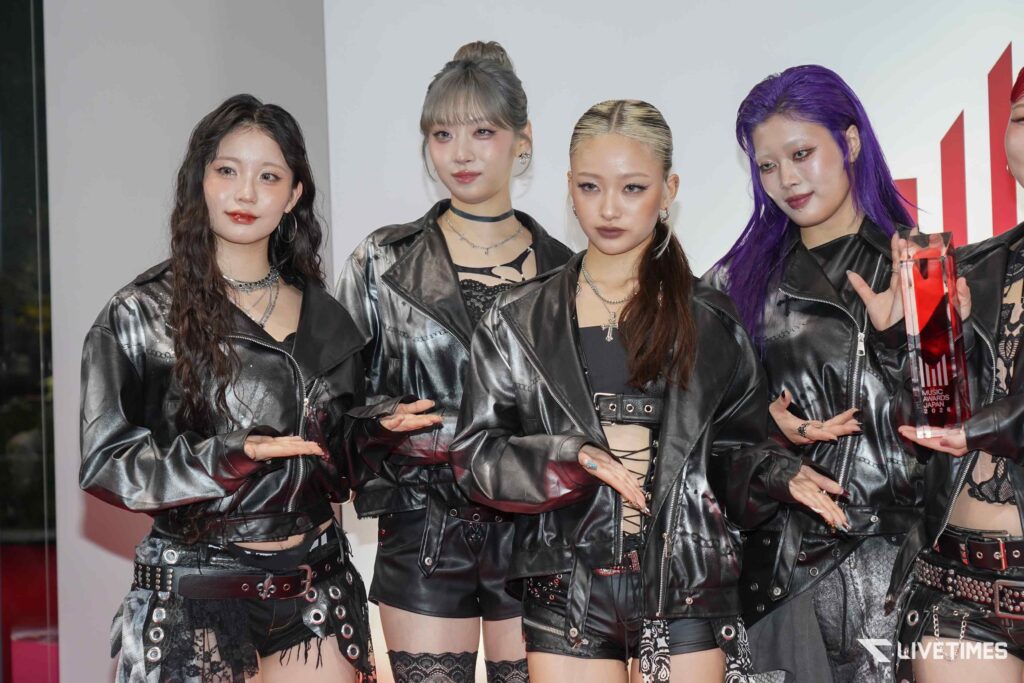 Four women in black leather outfits posing at an awards event, one holding a red crystal trophy on the right.