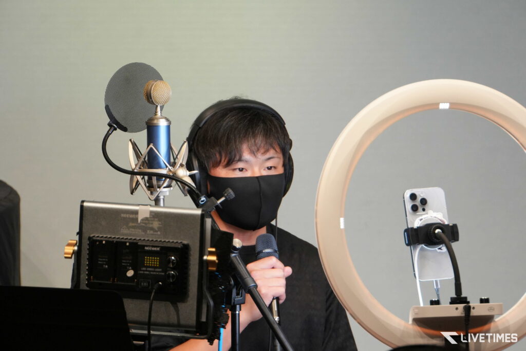 Person wearing a black mask and headphones speaks into a microphone in a recording studio, with a pop filter and a large ring light nearby.